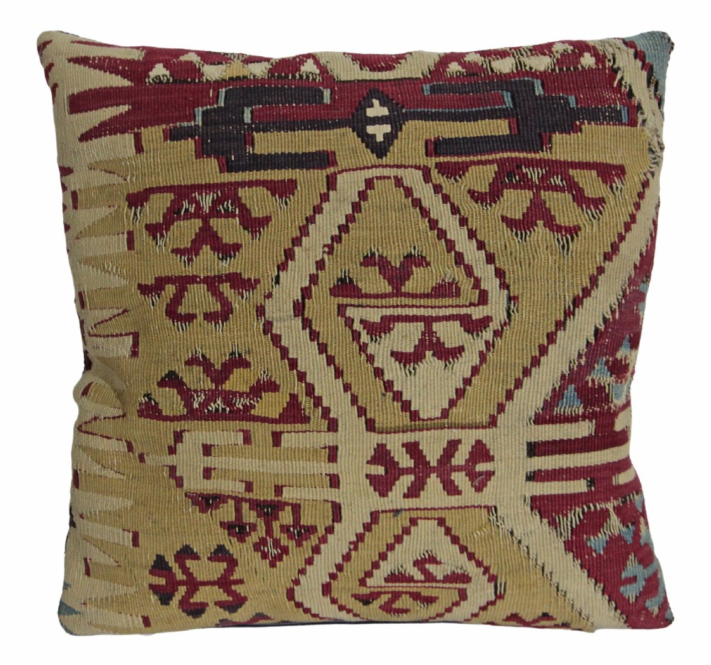 A square cushion crafted from flat-woven wool featuring geometric tribal motifs in beige, burgundy red, and dark blue hues.