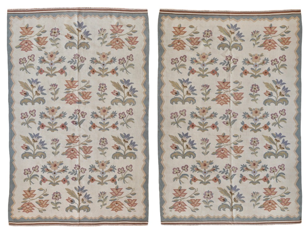 A matching pair of off-white hand-woven wool kilim rugs decorated with soft pink, blue, and sage floral motifs surrounded by a blue serrated border.