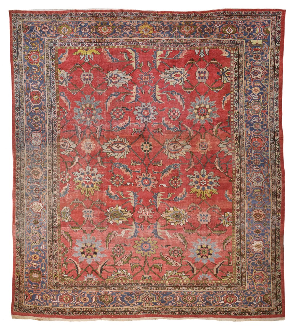 An antique hand-knotted Ziegler carpet featuring a terracotta red field with large-scale floral palmettes framed by a navy blue patterned border.