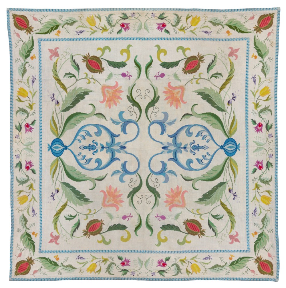 A square cream wool needlepoint rug featuring intricate green botanical scrollwork, pink blossoms, and pomegranate motifs within a decorative floral border.