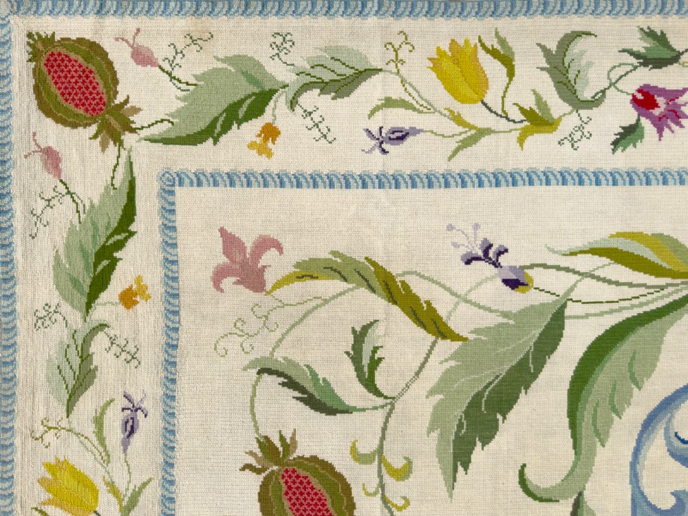 A square cream wool needlepoint rug featuring intricate green botanical scrollwork, pink blossoms, and pomegranate motifs within a decorative floral border. View VII