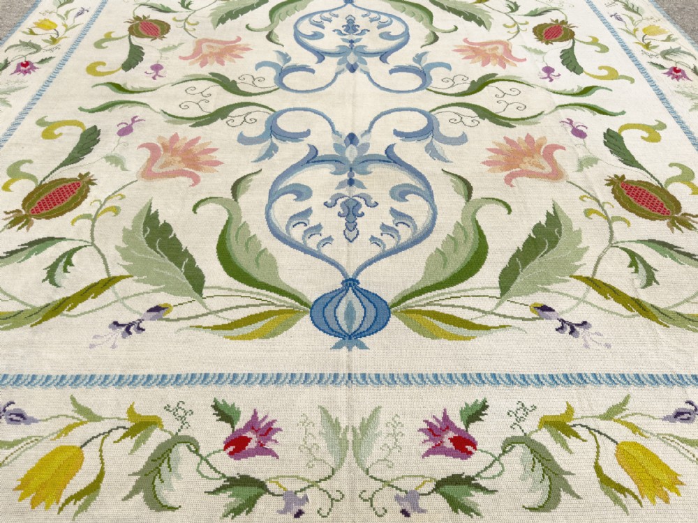 A square cream wool needlepoint rug featuring intricate green botanical scrollwork, pink blossoms, and pomegranate motifs within a decorative floral border. View IX