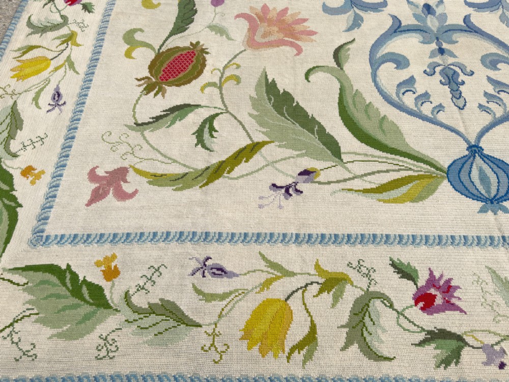A square cream wool needlepoint rug featuring intricate green botanical scrollwork, pink blossoms, and pomegranate motifs within a decorative floral border. View X