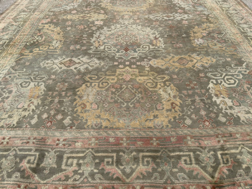 An antique hand-knotted Ushak wool carpet featuring a muted grey-green field adorned with central medallions, floral motifs, and multi-layered geometric borders in soft terracotta, pink, and gold tones. View V