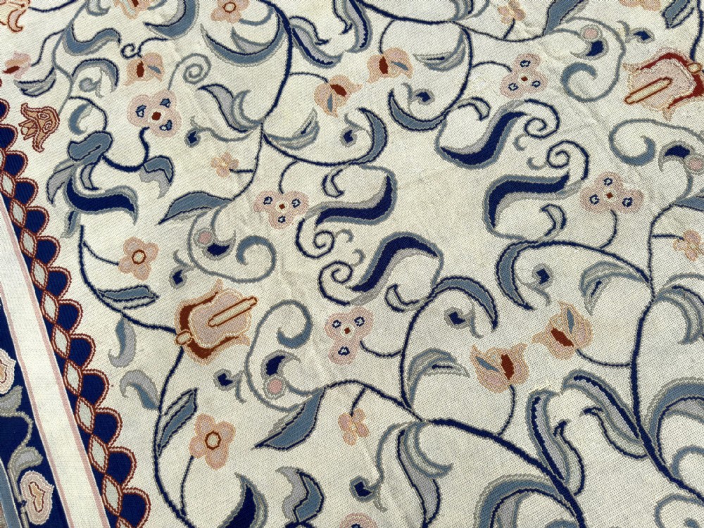 A rectangular Portuguese needlepoint wool rug featuring symmetrical floral scrollwork in blue and terracotta tones against an off-white field. View III