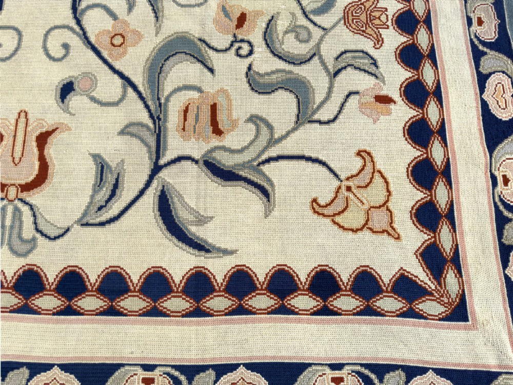 A rectangular Portuguese needlepoint wool rug featuring symmetrical floral scrollwork in blue and terracotta tones against an off-white field. View V
