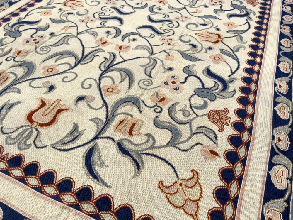A rectangular Portuguese needlepoint wool rug featuring symmetrical floral scrollwork in blue and terracotta tones against an off-white field. View VII