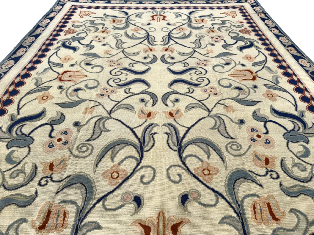 A rectangular Portuguese needlepoint wool rug featuring symmetrical floral scrollwork in blue and terracotta tones against an off-white field. View IX