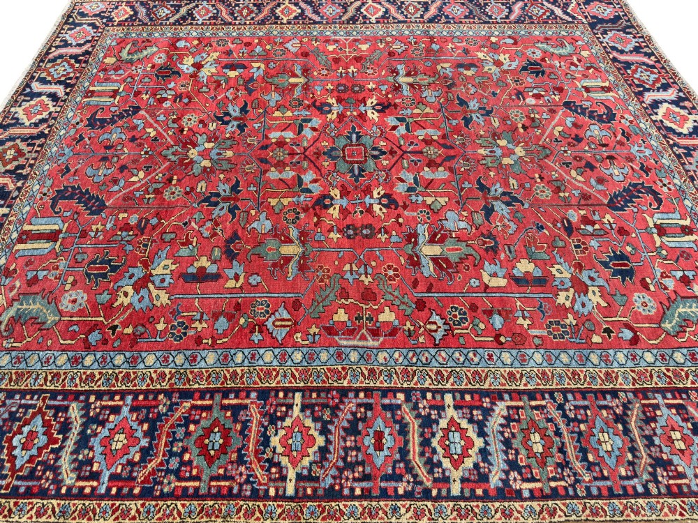 An antique hand-knotted Persian Heriz wool carpet featuring a central geometric medallion, detailed angular floral motifs on a terracotta red field, and a dark navy blue outer border. View III