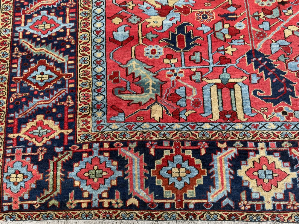 An antique hand-knotted Persian Heriz wool carpet featuring a central geometric medallion, detailed angular floral motifs on a terracotta red field, and a dark navy blue outer border. View IV