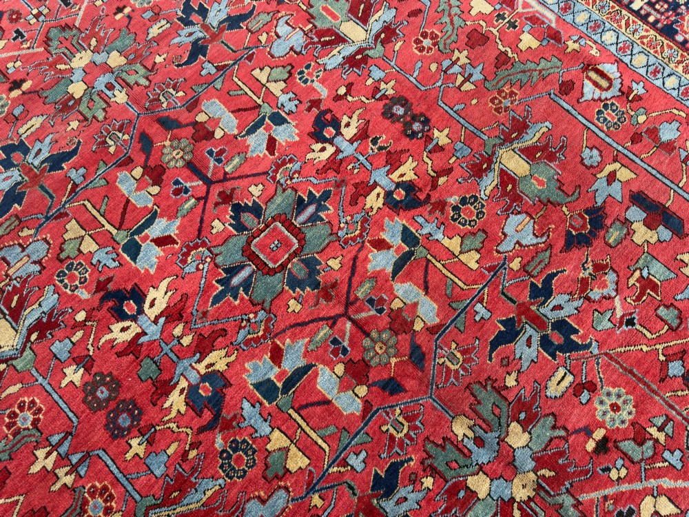An antique hand-knotted Persian Heriz wool carpet featuring a central geometric medallion, detailed angular floral motifs on a terracotta red field, and a dark navy blue outer border. View V