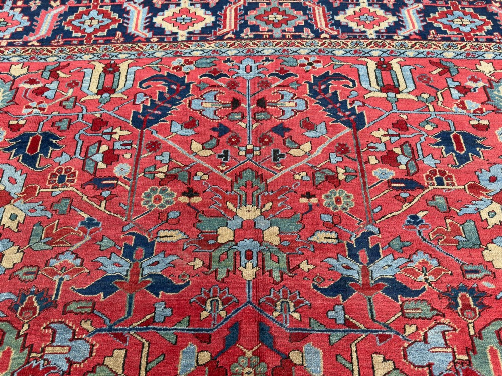 An antique hand-knotted Persian Heriz wool carpet featuring a central geometric medallion, detailed angular floral motifs on a terracotta red field, and a dark navy blue outer border. View VI