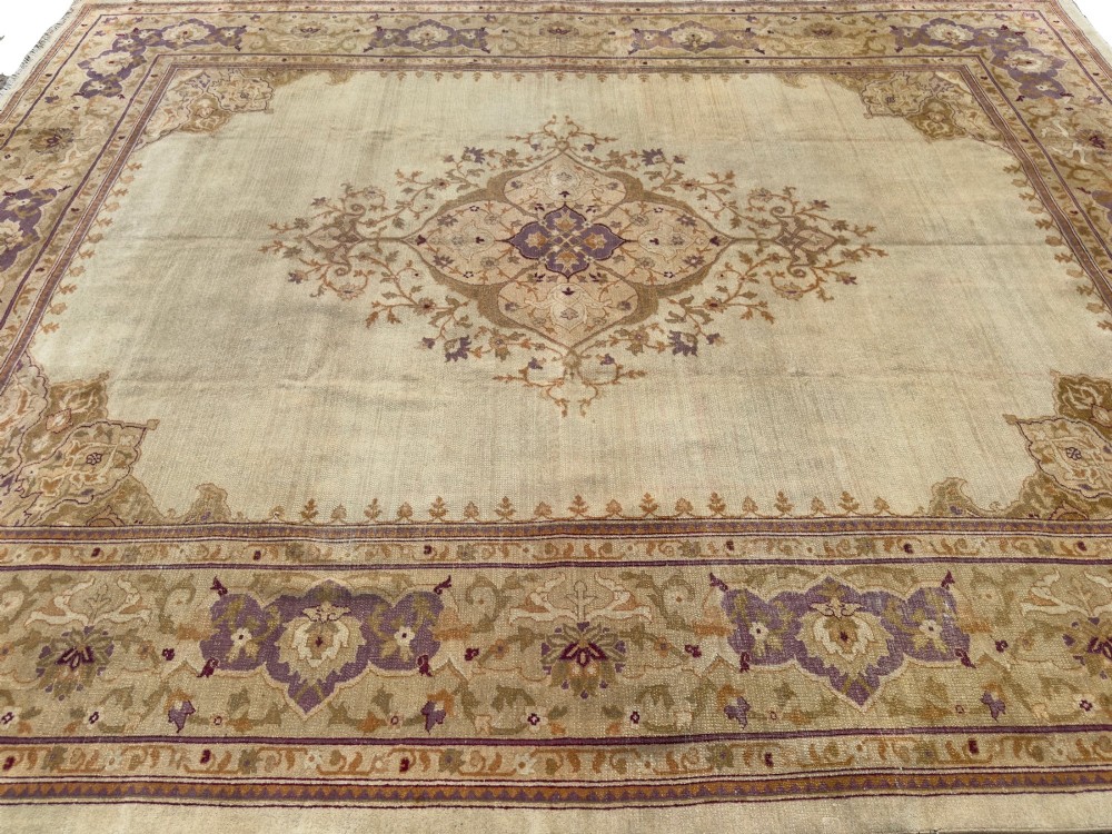 An antique hand-knotted rectangular wool carpet featuring a light cream field, a central floral medallion, corner spandrels, and a patterned border in shades of purple and ochre. View II