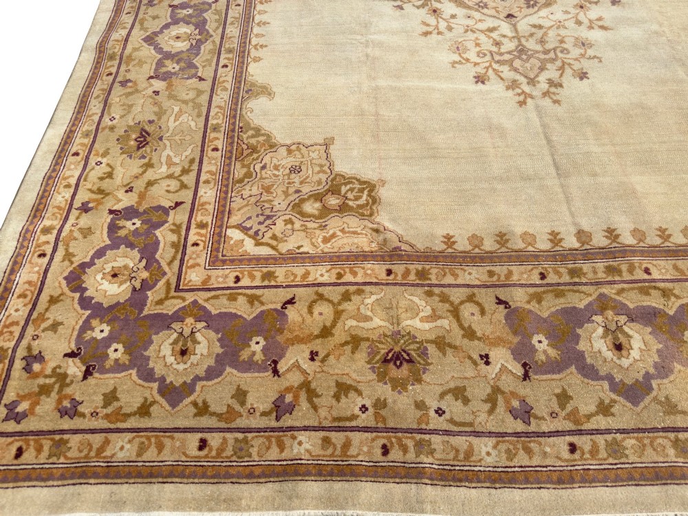 An antique hand-knotted rectangular wool carpet featuring a light cream field, a central floral medallion, corner spandrels, and a patterned border in shades of purple and ochre. View IV