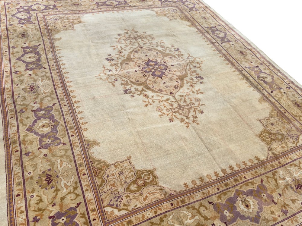 An antique hand-knotted rectangular wool carpet featuring a light cream field, a central floral medallion, corner spandrels, and a patterned border in shades of purple and ochre. View V