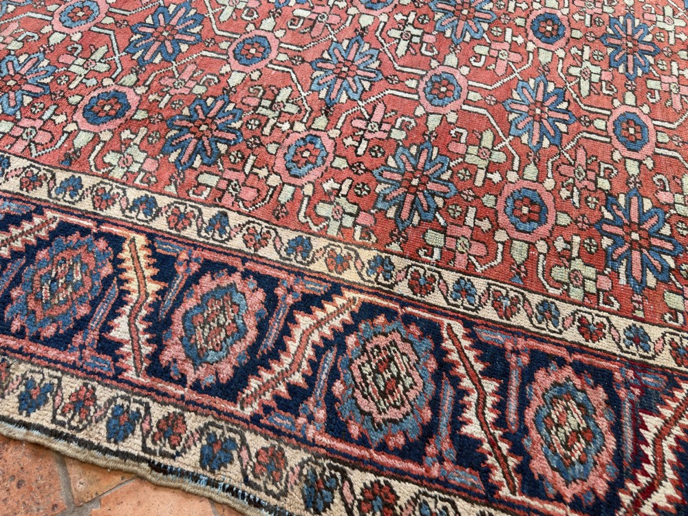 A hand-knotted antique Heriz wool carpet featuring a repeating geometric star motif on a terracotta red field framed by a wide navy blue patterned border. View III