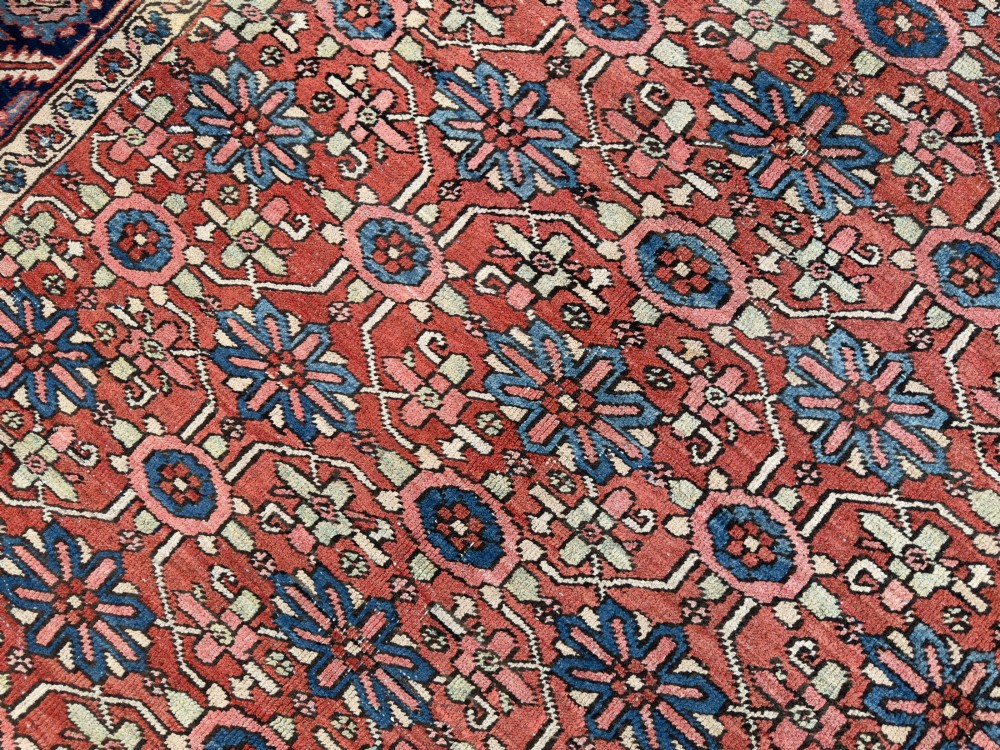 A hand-knotted antique Heriz wool carpet featuring a repeating geometric star motif on a terracotta red field framed by a wide navy blue patterned border. View IV