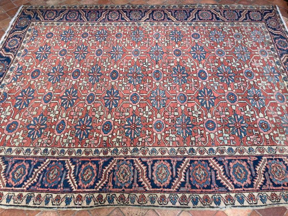 A hand-knotted antique Heriz wool carpet featuring a repeating geometric star motif on a terracotta red field framed by a wide navy blue patterned border. View V