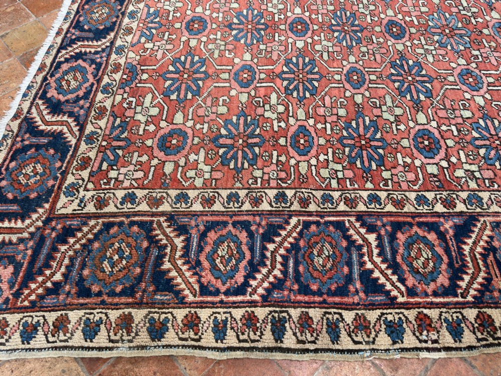 A hand-knotted antique Heriz wool carpet featuring a repeating geometric star motif on a terracotta red field framed by a wide navy blue patterned border. View VI