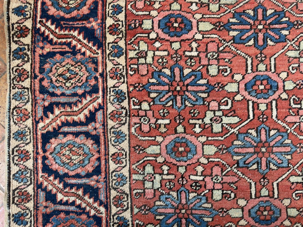 A hand-knotted antique Heriz wool carpet featuring a repeating geometric star motif on a terracotta red field framed by a wide navy blue patterned border. View VII