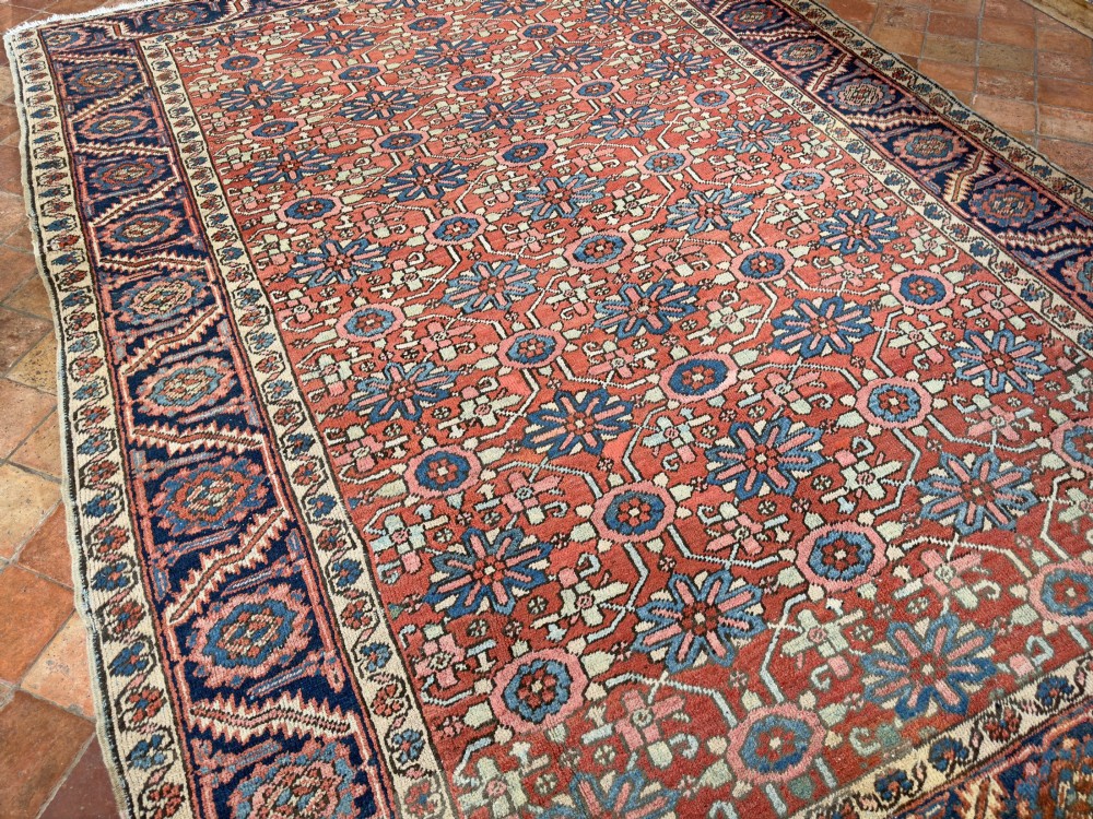 A hand-knotted antique Heriz wool carpet featuring a repeating geometric star motif on a terracotta red field framed by a wide navy blue patterned border. View VIII