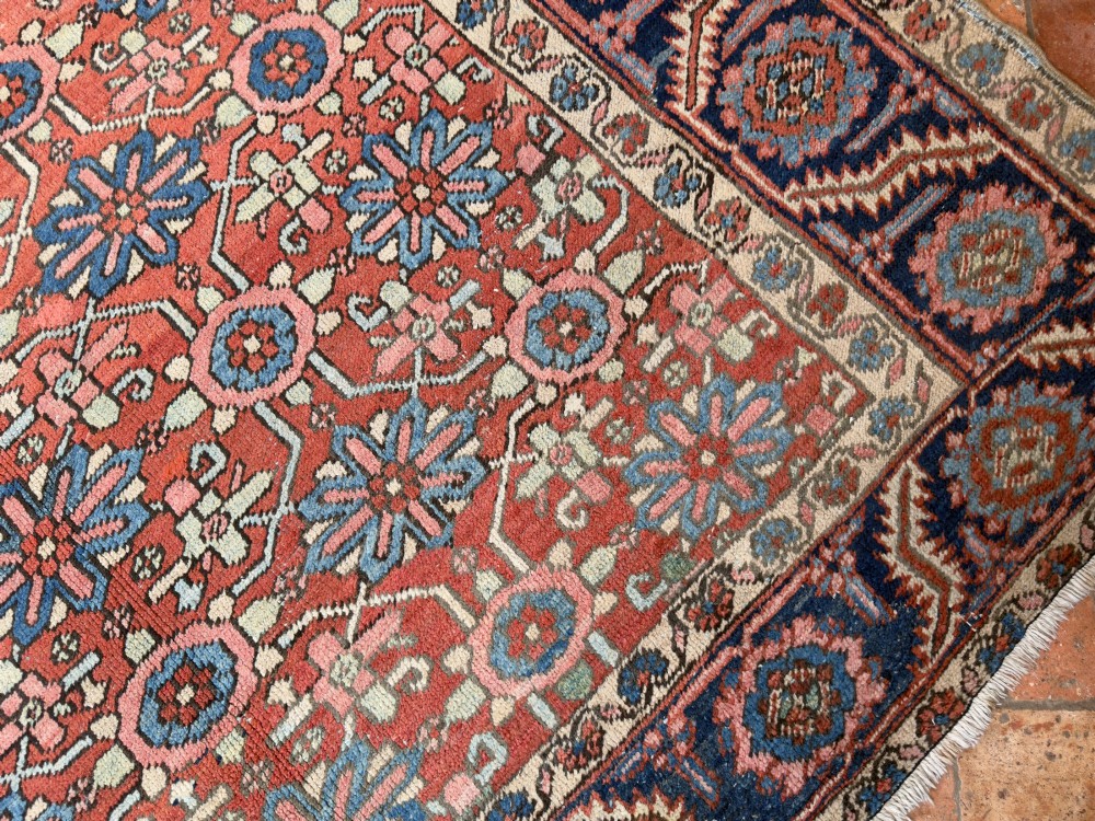 A hand-knotted antique Heriz wool carpet featuring a repeating geometric star motif on a terracotta red field framed by a wide navy blue patterned border. View X