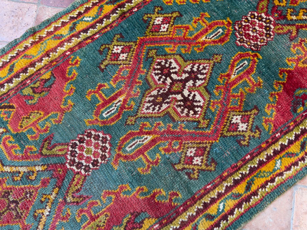 An antique hand-knotted wool runner rug featuring geometric tribal medallions in red, yellow, and orange on a vibrant teal background. View V
