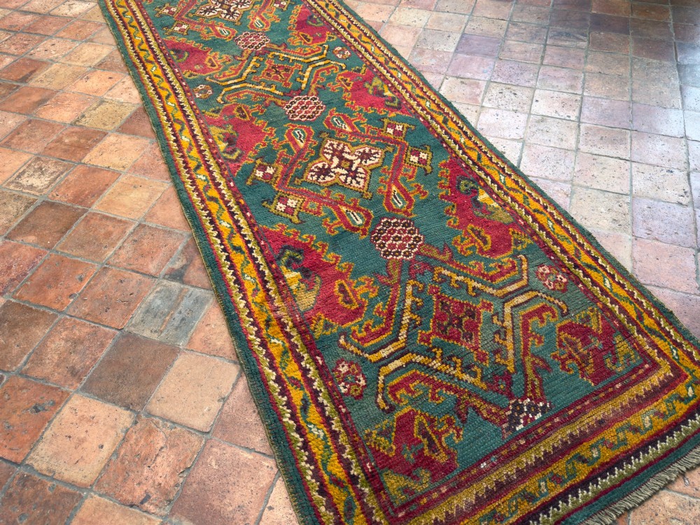 An antique hand-knotted wool runner rug featuring geometric tribal medallions in red, yellow, and orange on a vibrant teal background. View VI