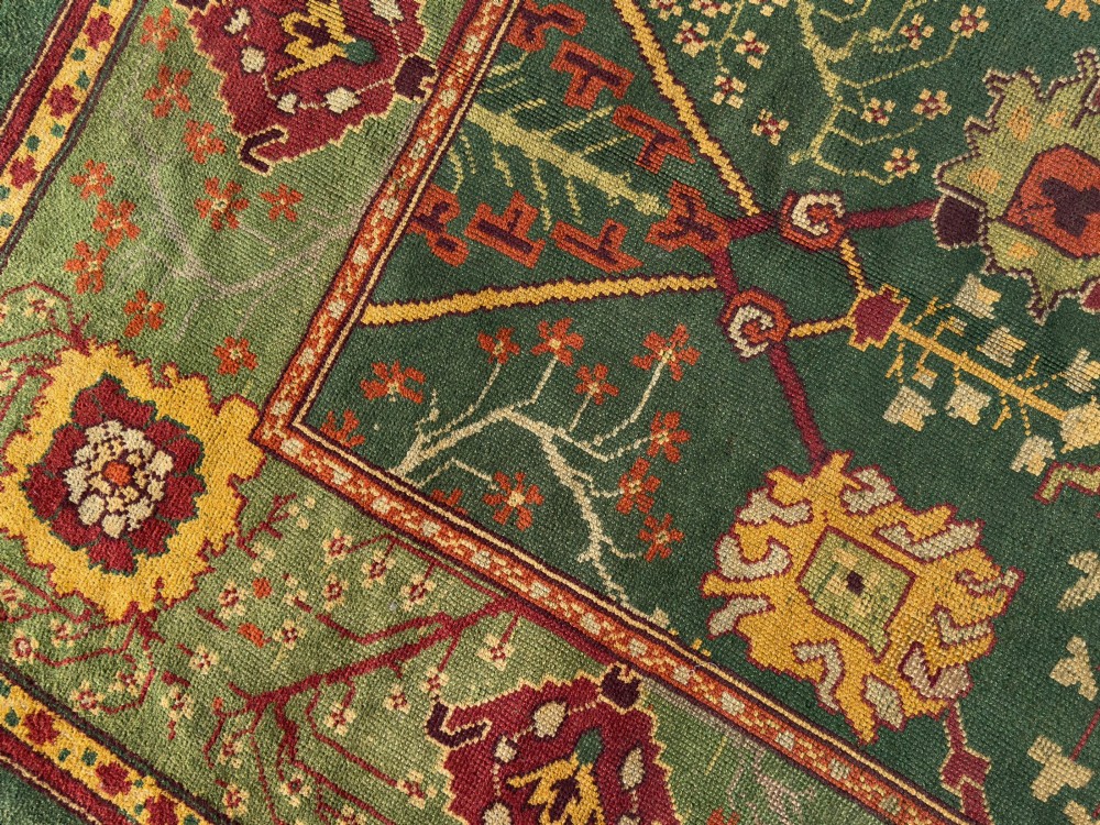 An antique Arts and Crafts hand-knotted wool Donegal carpet featuring a green field filled with stylised red and yellow floral motifs and a decorative geometric border. View X