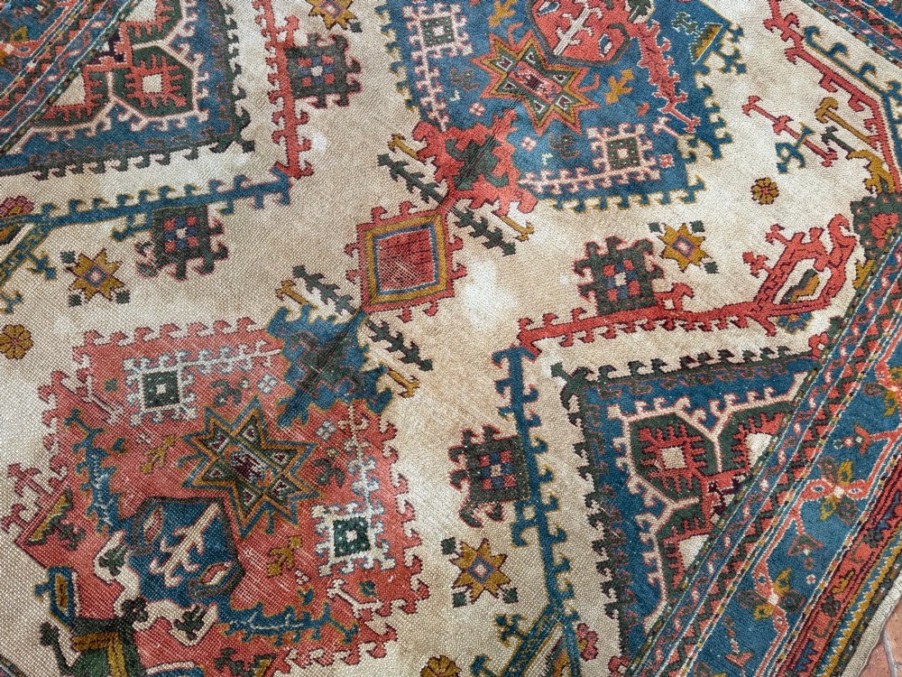 A rectangular hand-knotted wool Ushak carpet featuring central geometric diamond medallions in terracotta red and blue set against a cream field with framed borders. View III