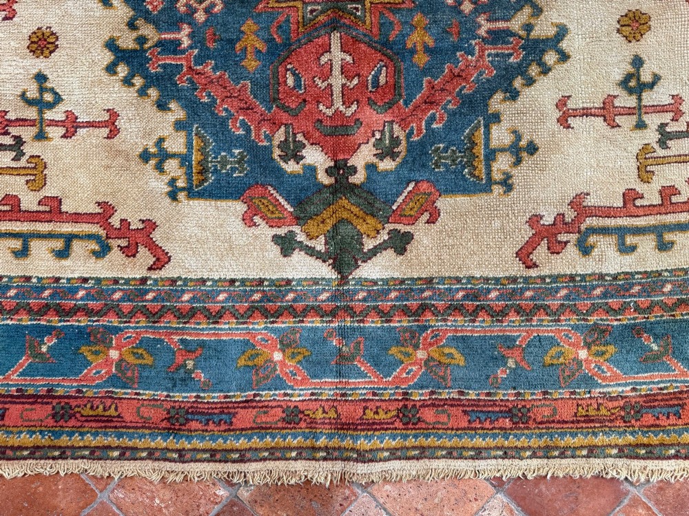A rectangular hand-knotted wool Ushak carpet featuring central geometric diamond medallions in terracotta red and blue set against a cream field with framed borders. View IV