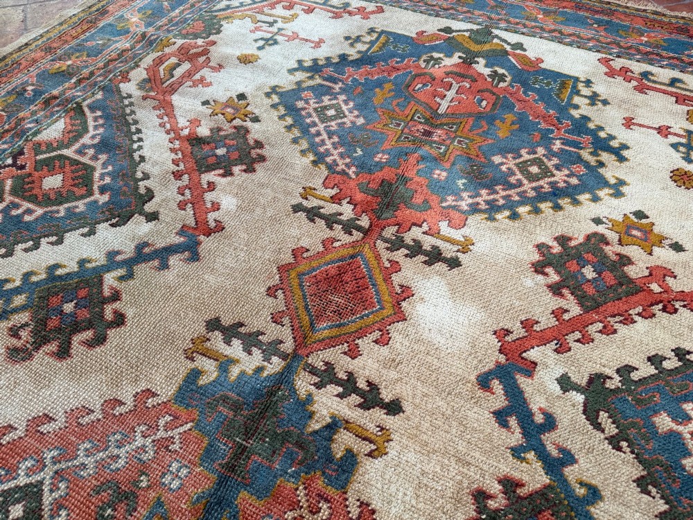 A rectangular hand-knotted wool Ushak carpet featuring central geometric diamond medallions in terracotta red and blue set against a cream field with framed borders. View V