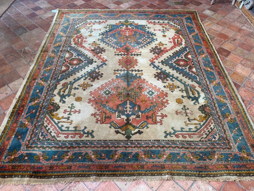 A rectangular hand-knotted wool Ushak carpet featuring central geometric diamond medallions in terracotta red and blue set against a cream field with framed borders. View VI