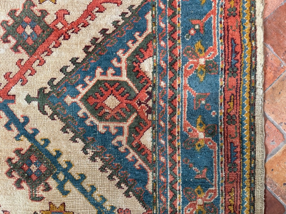 A rectangular hand-knotted wool Ushak carpet featuring central geometric diamond medallions in terracotta red and blue set against a cream field with framed borders. View VII