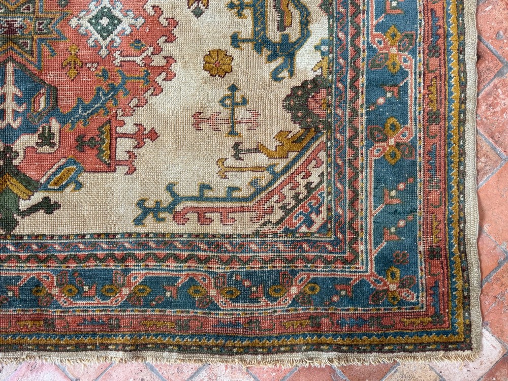 A rectangular hand-knotted wool Ushak carpet featuring central geometric diamond medallions in terracotta red and blue set against a cream field with framed borders. View VIII