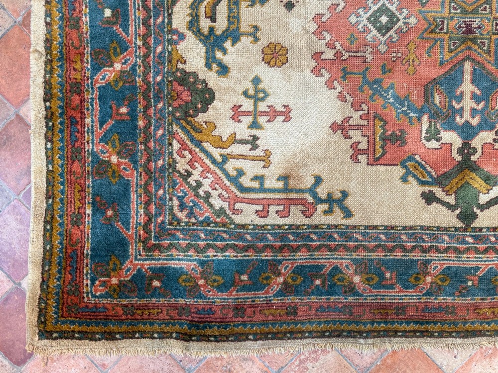 A rectangular hand-knotted wool Ushak carpet featuring central geometric diamond medallions in terracotta red and blue set against a cream field with framed borders. View IX