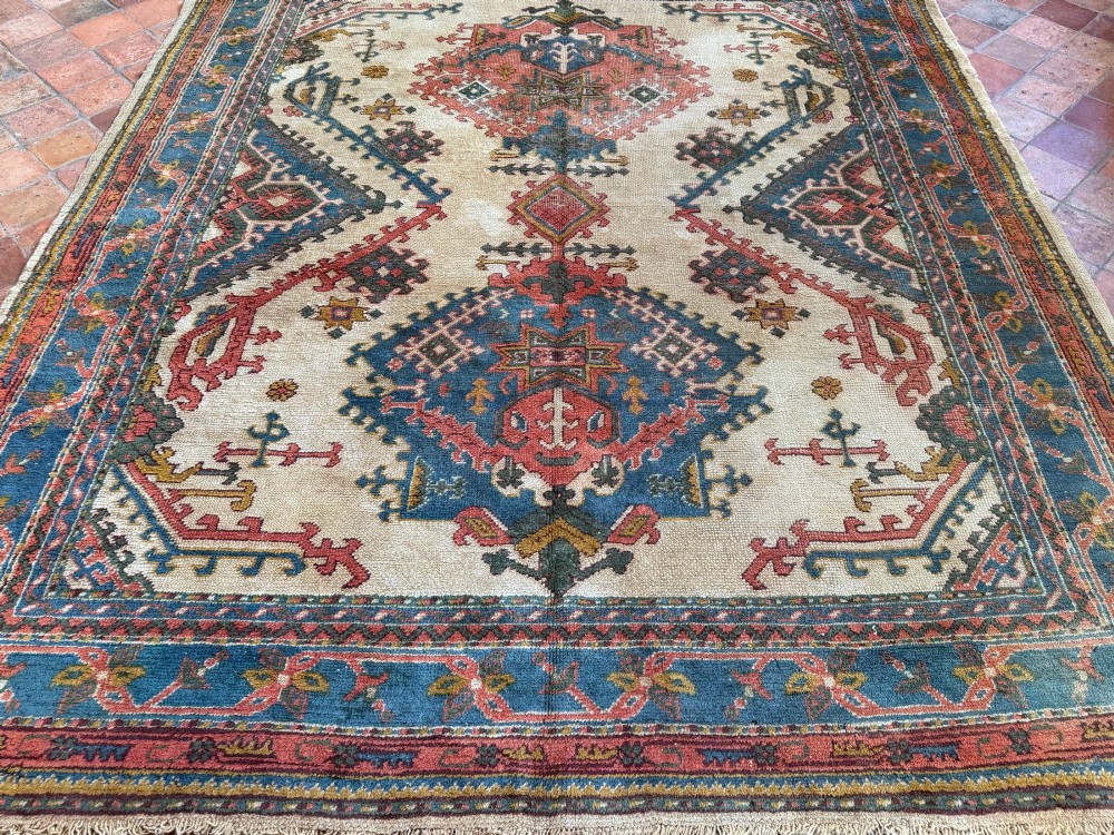A rectangular hand-knotted wool Ushak carpet featuring central geometric diamond medallions in terracotta red and blue set against a cream field with framed borders. View X