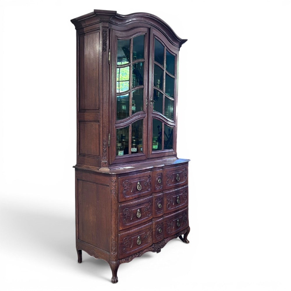 An 18th-century French dark oak two-piece cabinet featuring a carved commode base with brass handles beneath an arched glazed vitrine top.