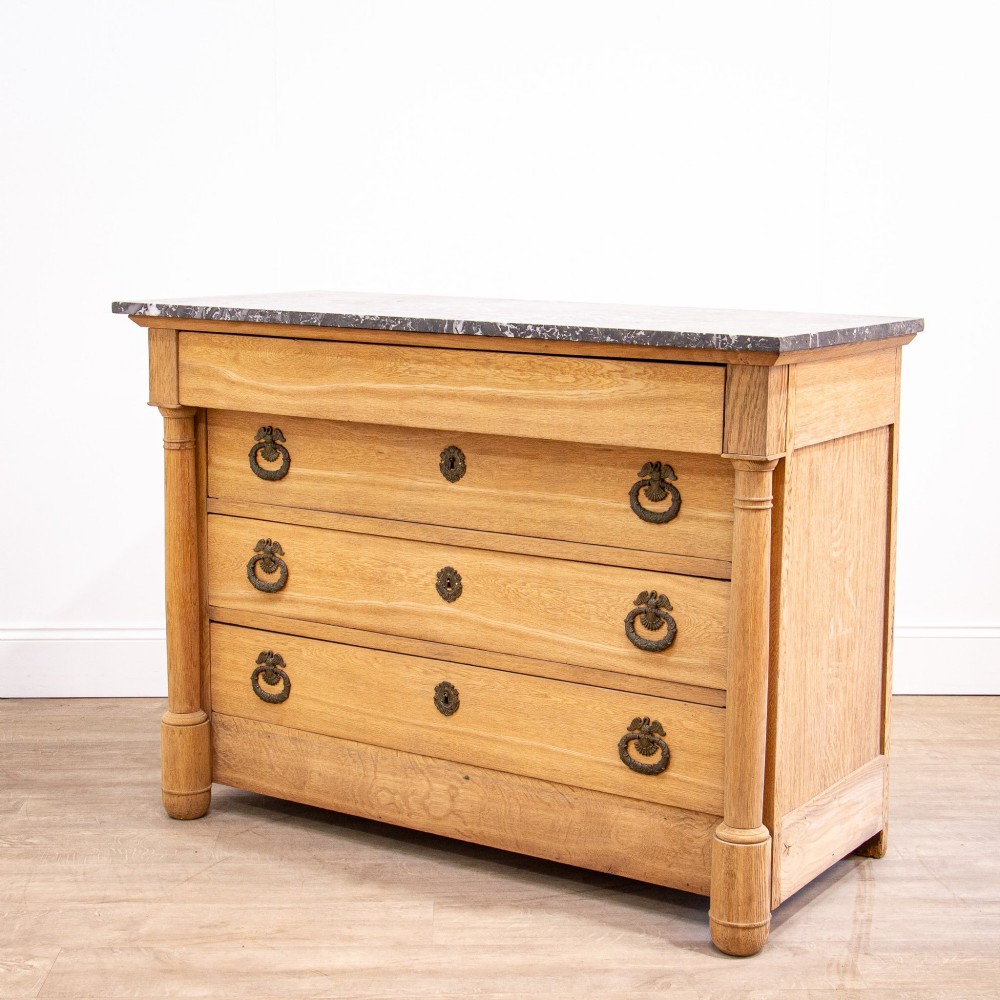 A nineteenth-century bleached oak French Empire commode featuring four drawers, flanking columns, brass ring pull handles, and a dark grey marble top.