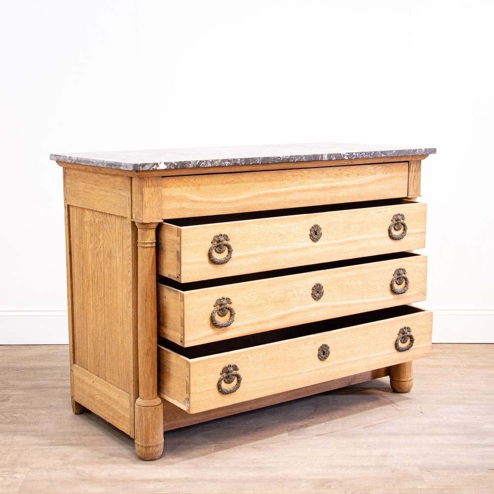 A nineteenth-century bleached oak French Empire commode featuring four drawers, flanking columns, brass ring pull handles, and a dark grey marble top. View II