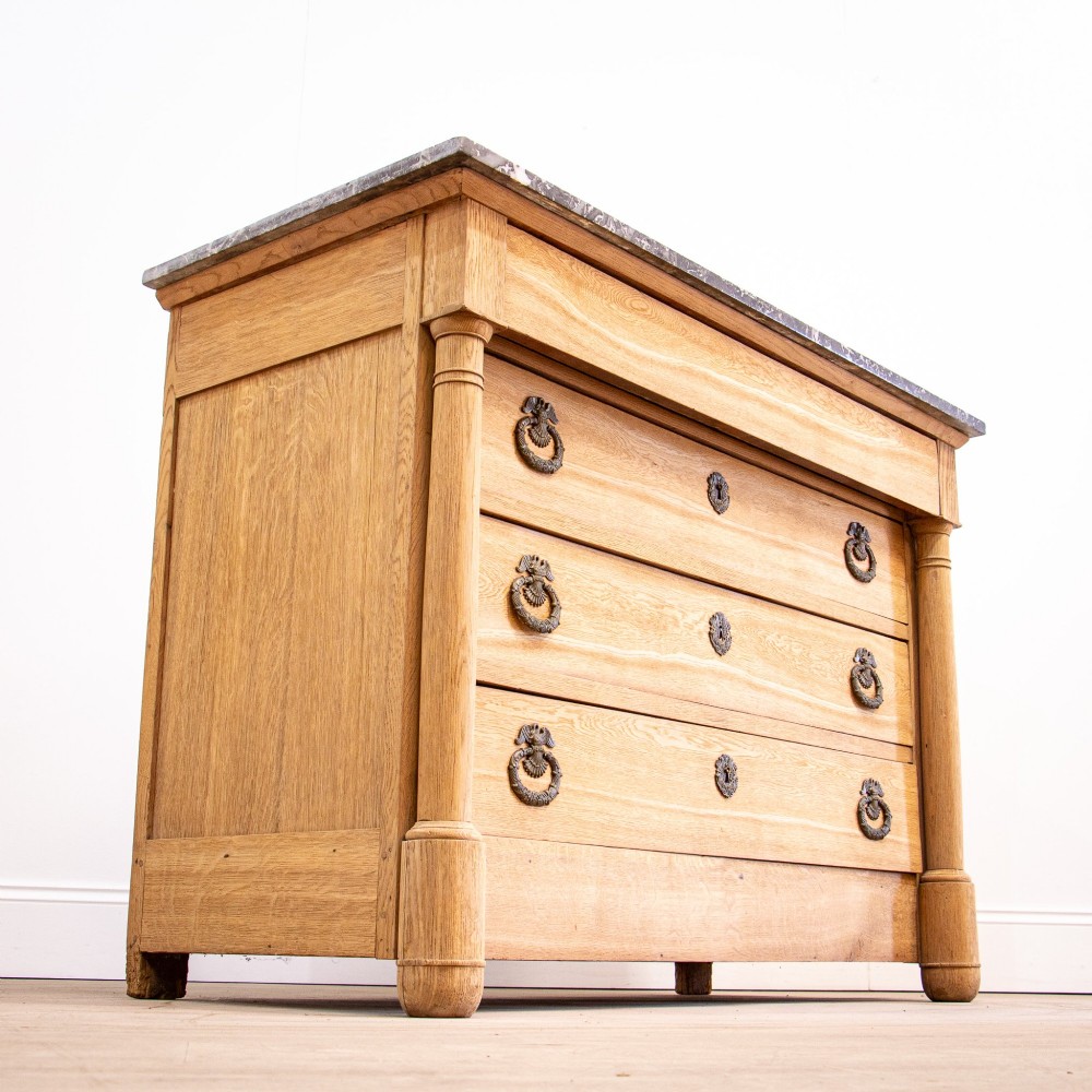 A nineteenth-century bleached oak French Empire commode featuring four drawers, flanking columns, brass ring pull handles, and a dark grey marble top. View V