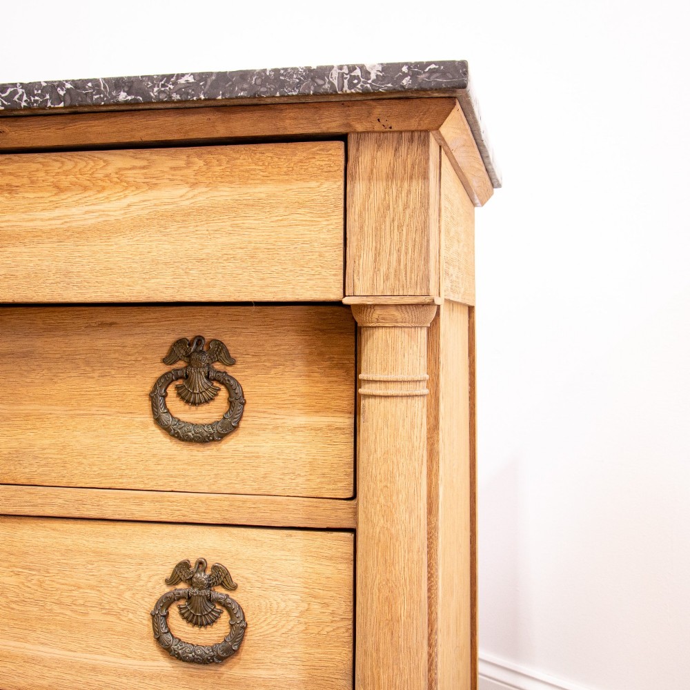 A nineteenth-century bleached oak French Empire commode featuring four drawers, flanking columns, brass ring pull handles, and a dark grey marble top. View X