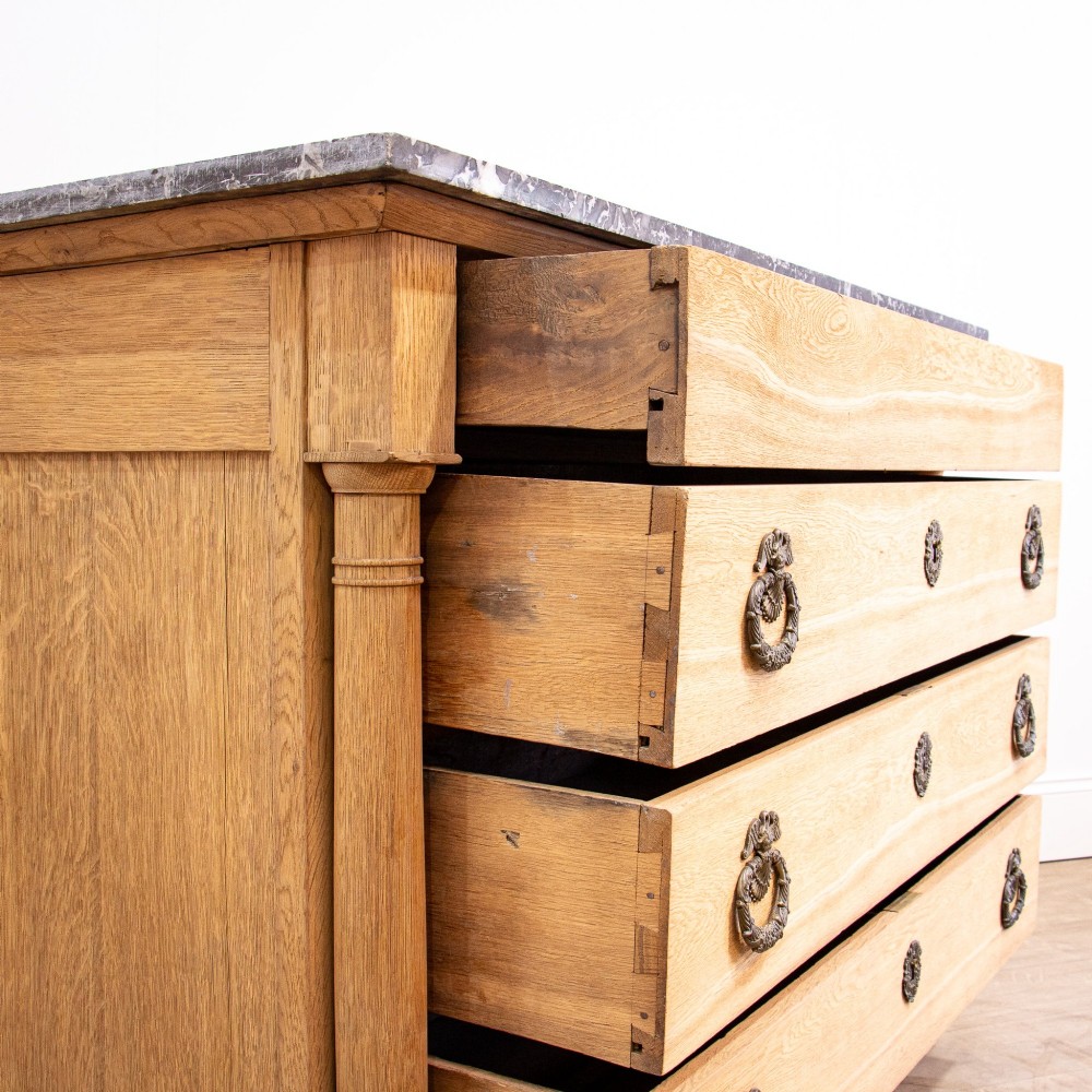 A nineteenth-century bleached oak French Empire commode featuring four drawers, flanking columns, brass ring pull handles, and a dark grey marble top. View XII