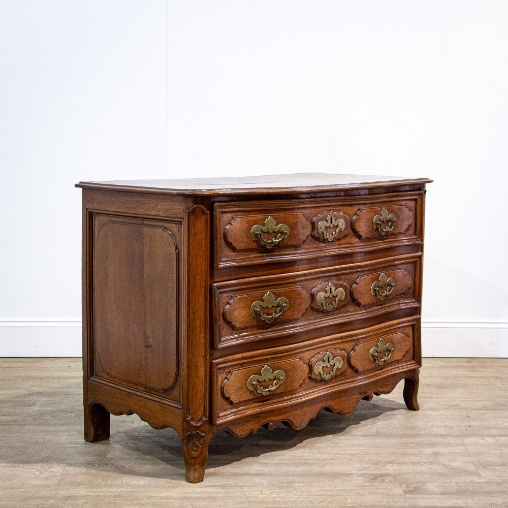An 18th-century French Louis XV oak commode featuring three serpentine drawers with carved moulded panels, ornate brass handles, and carved cabriole legs.