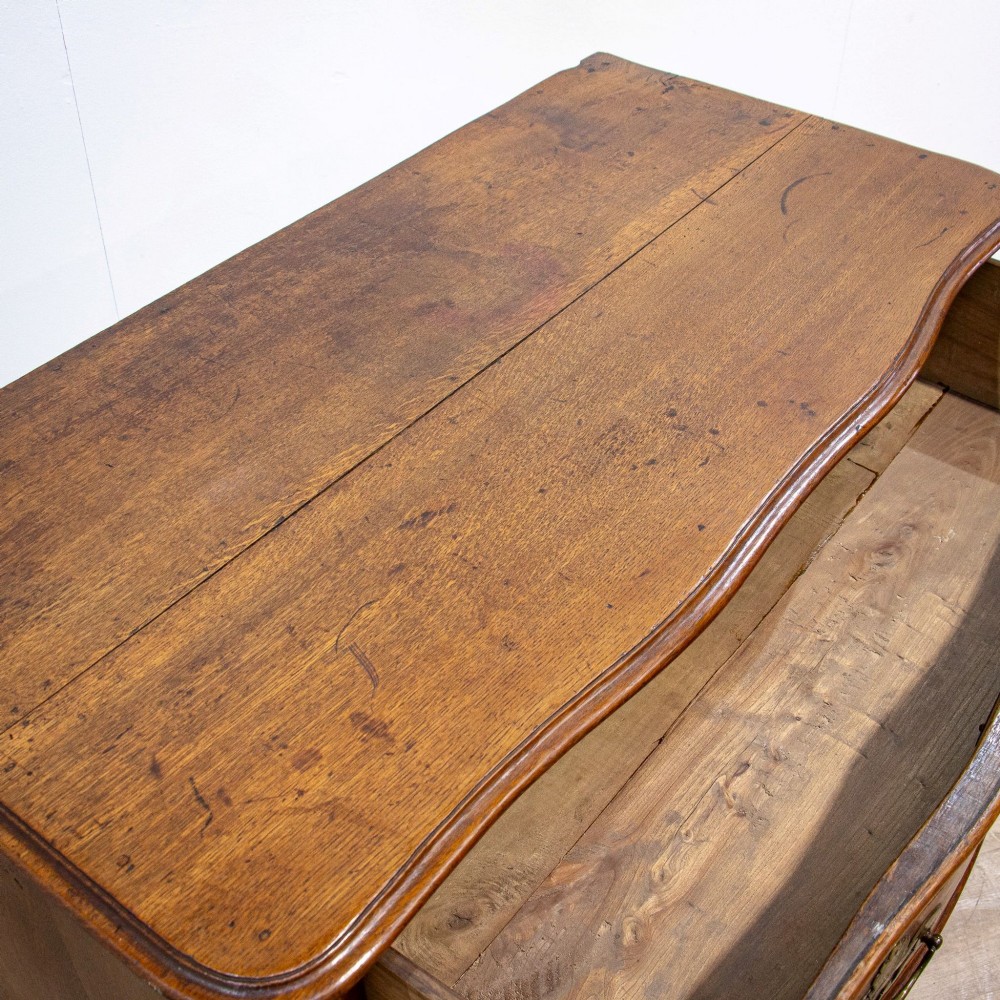 An 18th-century French Louis XV oak commode featuring three serpentine drawers with carved moulded panels, ornate brass handles, and carved cabriole legs. View IV