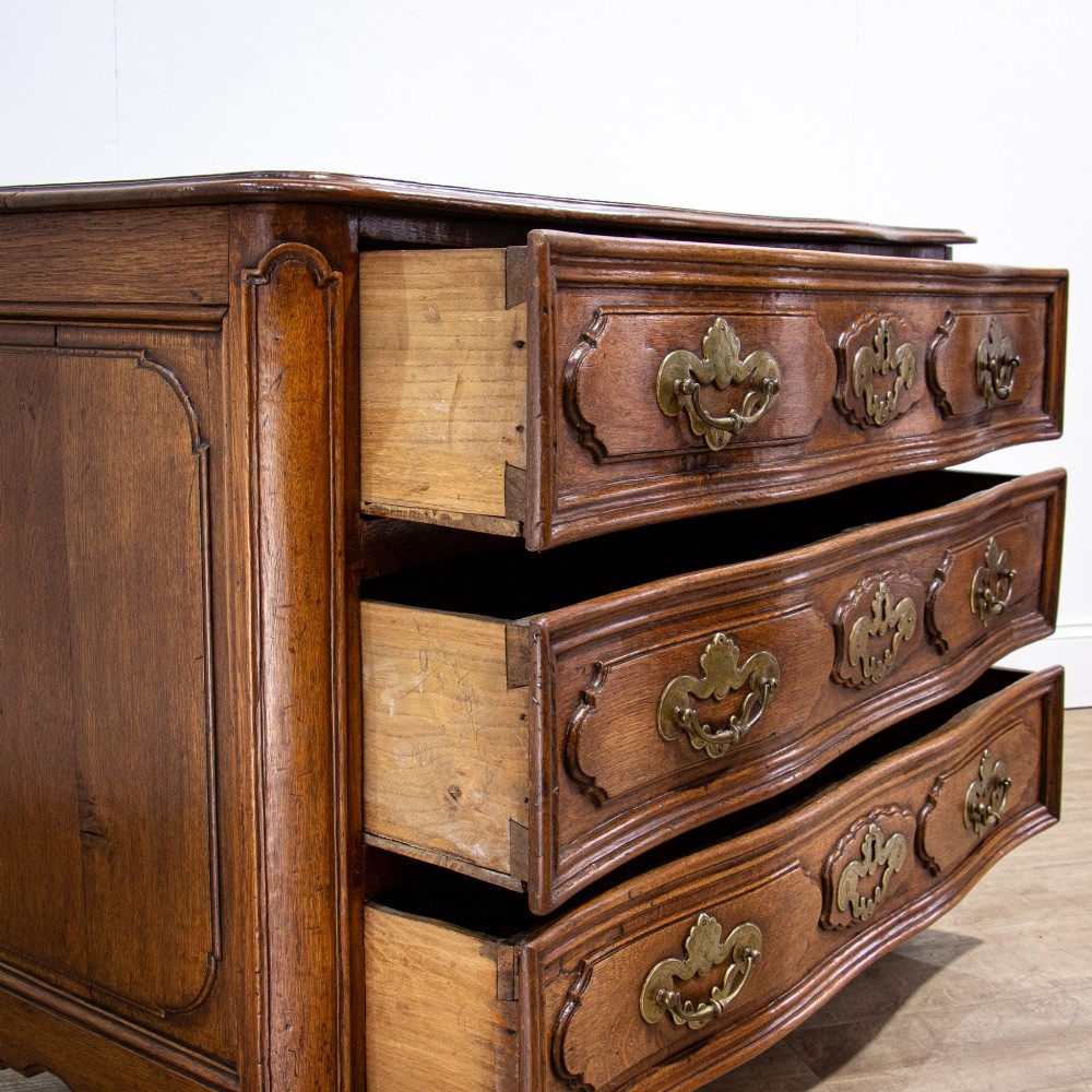 An 18th-century French Louis XV oak commode featuring three serpentine drawers with carved moulded panels, ornate brass handles, and carved cabriole legs. View IX