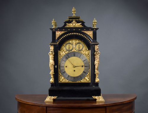 fig 1. A Victorian ebonised bracket clock featuring an arched black wooden case, gilded brass caryatids, a gold spandrel dial with Roman numerals, and top flame finials.