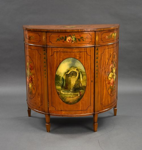 fig 1. An Edwardian demi-lune satinwood cabinet featuring hand-painted floral and landscape medallions on two doors and tapered legs.