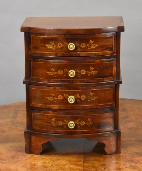 fig 1. A miniature warm brown wooden chest of drawers featuring a bow front, four drawers with floral marquetry inlay, and central brass knobs.