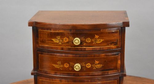 fig 2. A miniature warm brown wooden chest of drawers featuring a bow front, four drawers with floral marquetry inlay, and central brass knobs.